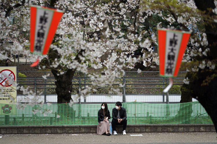 Мъж и жена със защитни маски седят на прочутата улица на цъфтящите сакури в парк Уено в Токио, петък, 27 март 2020 г. Улицата беше затворена по-късно, като предпазна мярка срещу коронавируса.